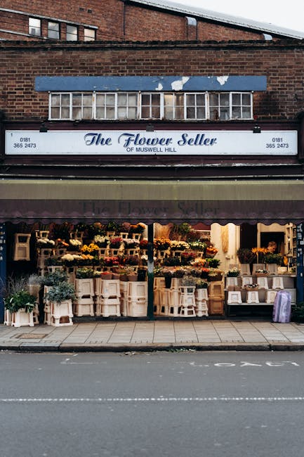 The exterior of 'The Flower Seller of Muswell Hill', showcasing large display windows filled with an array of vibrant, fresh flowers arranged in bouquets, vases, and floral displays. The shop features