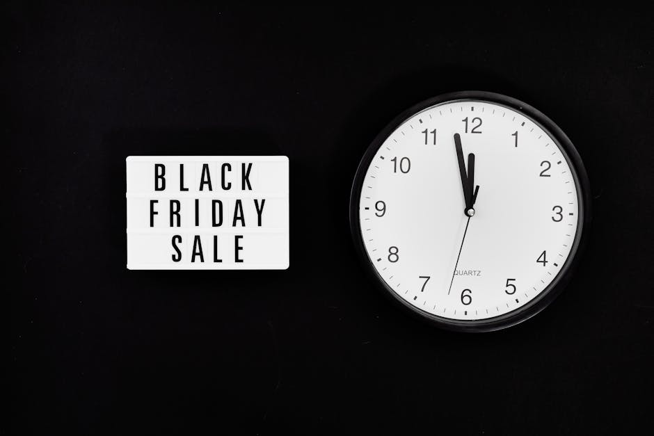 A white lightbox displaying black text reading 'BLACK FRIDAY SALE' is positioned on a dark surface to the left of a round, white-faced quartz clock with black hour, minute, and second hands. The clock
