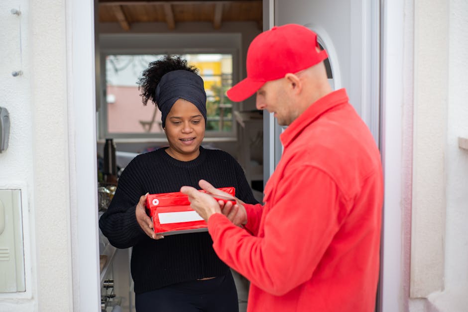 A flower delivery courier in a red uniform and cap hands a red box to a woman with dark curly hair wrapped in a black headscarf, standing at her doorstep with a neutral expression. The woman is receiv