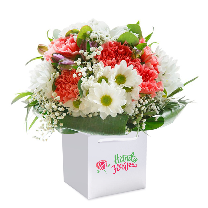 An enchanting floral arrangement rises upright from a sleek white gift bag, exemplifying the creative heart of Mile End's floral community. This premium bouquet brings together lavish coral and peach carnations, their signature frilly, layered petals radiating warmth and energy. Bright white daisies with lively lime-green centres pop intermittently among the more subdued tones, evoking the cheerful energy of Victoria Park on a sunny day. Ethereal sprays of white baby's breath thread delicately between the larger blooms, adding lightness and dynamic movement to the design. Nestled quietly to the left are purple-tinged buds-likely alstroemeria-hinting at growth and renewal. Robust arcs of glossy, emerald foliage anchor the arrangement, fanning out with the structured elegance of the trees lining Mile End Road. The clean, sturdy white bag features matching ribbon handles that speak of thoughtful, practical presentation-ideal for a seamless local delivery. Arranged in a full, rounded silhouette, each flower sits at its best angle, broadcasting freshness and abundance. The visual poetry of this bouquet, a blend of colour, texture, and form, makes it perfectly suited for a heartfelt gift-be it birthday, anniversary, or simply conveying care-delivered with pride throughout Mile End.