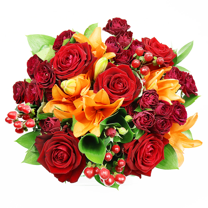 Captured from above on a crisp white surface, this abundant bouquet presents a rich tapestry of reds, orange and green in a compact, circular form. Several large deep red roses dominate the eye, their velvety petals unfolding in classic spirals and creating a sense of timeless romance. Bright orange Asiatic lilies are threaded through the roses, their trumpet-shaped petals and visible stamens offering bursts of warmth; some lilies stand fully open, declaring themselves, while other buds remain gracefully furled, suggesting freshness and an evolving bloom. Interwoven are tight clusters of deep burgundy spray roses whose miniature heads create velvety shadow and luxurious texture, giving the arrangement a layered, jewel-like quality. Scattered throughout are glossy hypericum berries-small, round and ruby red-that add playful punctuation and tactile contrast to the soft petals. The green foliage, broad and lustrous like ruscus or salal, peeks through the massed flowers, providing a cool, grounding backdrop that lifts the composition's palette. The lighting is mild and even, revealing the interplay of matte and glossy surfaces: the rose velvet, the lily satin, the berry sheen and the leaf gloss. Imagine holding this bouquet on an overcast morning after a quick walk past Burdett Road shops to the arranging table-its scent blends the heady sweetness of roses with a fresh, spicy hint from the lilies. This design feels celebratory and intimate at once, suited to anniversaries, romantic gestures, or as a warming statement piece for a home in Mile End.