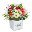 This carefully crafted bouquet exudes an upbeat Mile End vibrancy, lovingly assembled in a gleaming square white gift bag with smart ribbon handles. A lively medley of coral, peach, and lively red carnations unfolds in velvety ruffles, their textures inviting touch. Cheery white daisies, each shining with a sunlit yellow-green centre, dot the arrangement, adding pure joy reminiscent of picnics in local parks. Sprays of delicate, airy baby's breath drift through, softening the scene like morning mist. At the periphery, a secret cluster of purple-hued buds hints at surprises yet to bloom. Encircling the flowers, broad arcs of deep, glossy green foliage provide sturdy rhythm and visual richness, grounding the bouquet in natural sophistication. The arrangement's perfect, rounded form and crisp lines set a tone of freshness and abundance, echoing the scenic pathways of Mile End. Delivered ready for any occasion-a birthday, an anniversary, or simply to spark delight in a loved one's home-this bouquet is a joyful tribute to the area's friendly community, its colours and scents a vibrant celebration of local floral artistry.